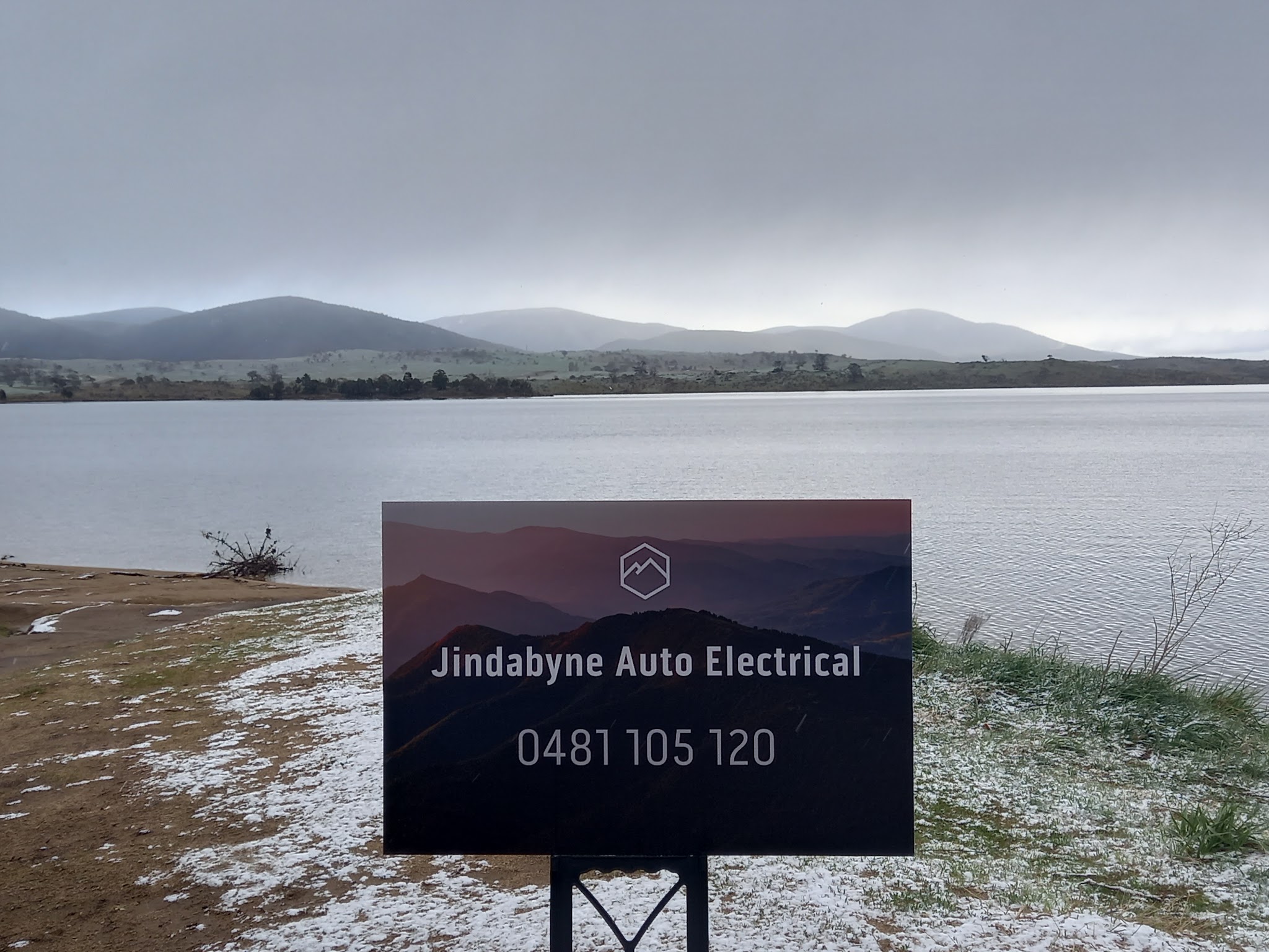 Jindabyne Auto Electrical sign board on the snow dusted shore of Lake Jindabyne, with the Snowy Mountains behind